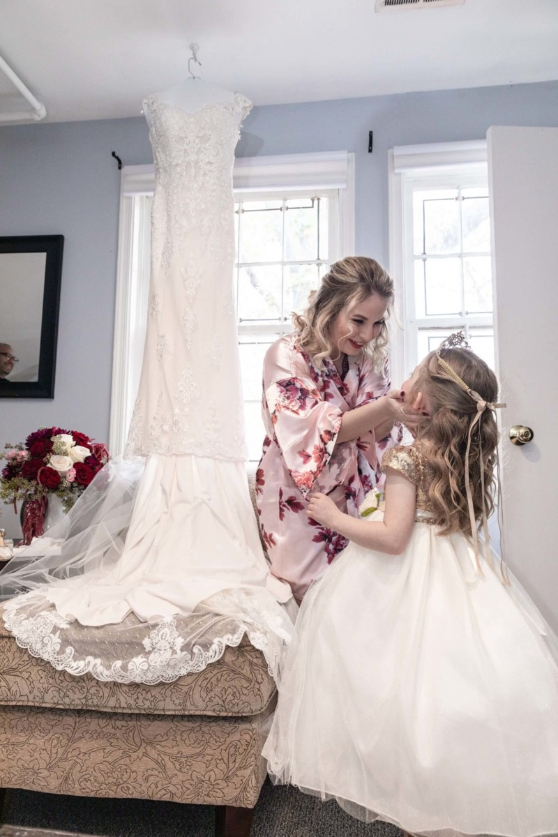 Shannon-James-Wedding-Low-Res-32 - TOMMY GATZ ENTERTAINMENT A woman in a floral robe and a young girl in a white dress share a moment beside a white bridal gown displayed on a chair.