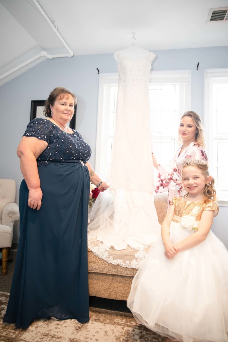 Shannon-James-Wedding-Low-Res-33 - TOMMY GATZ ENTERTAINMENT Three people pose indoors with a wedding dress hanging on a stand.