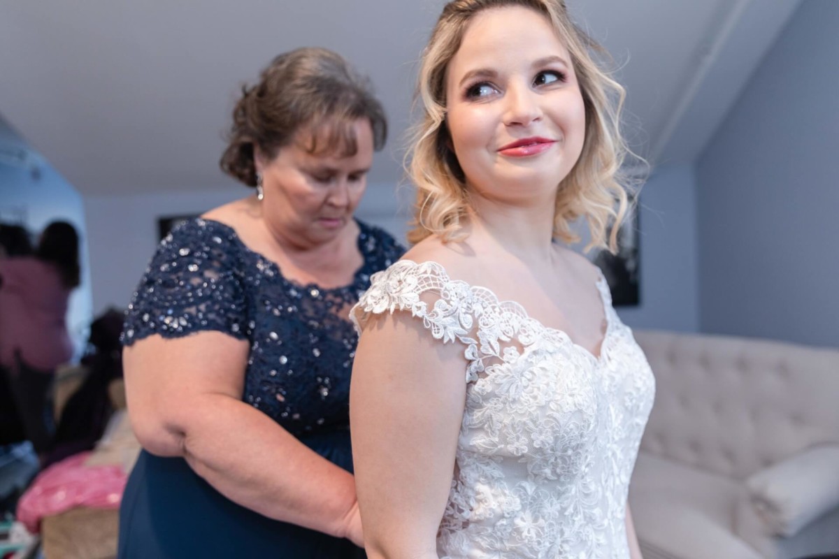 Shannon-James-Wedding-Low-Res-34 - TOMMY GATZ ENTERTAINMENT A woman in a lace wedding dress smiles as another woman helps her with the back of the dress in a room with a beige couch.