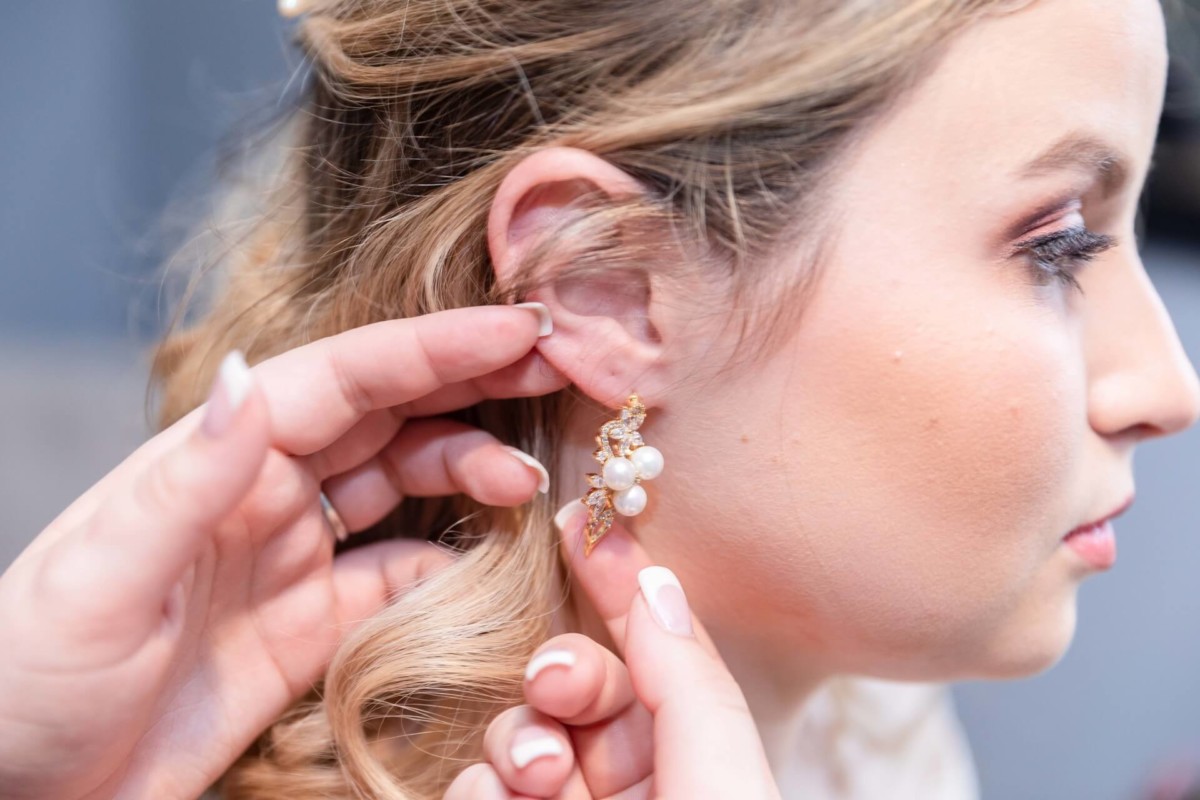 Shannon-James-Wedding-Low-Res-37 - TOMMY GATZ ENTERTAINMENT Person inserting a pearl and gold earring into a woman's ear. The woman's hair is styled in loose curls.