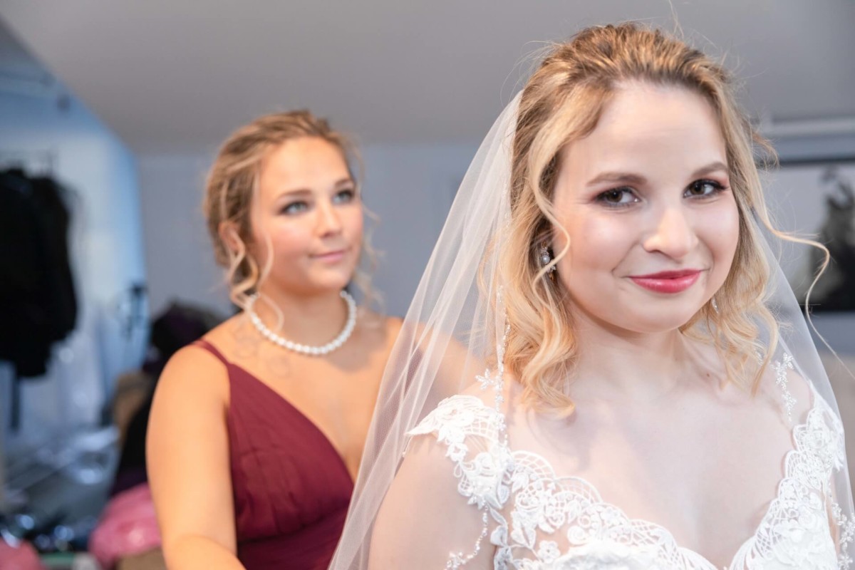 Shannon-James-Wedding-Low-Res-43 - TOMMY GATZ ENTERTAINMENT Bride smiling in a white dress and veil, standing in the foreground, with a bridesmaid in a maroon dress in the background.