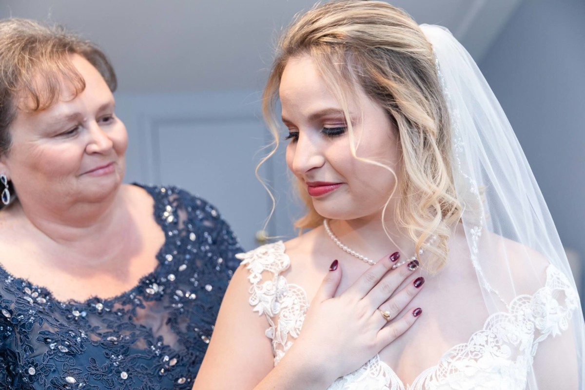 Shannon-James-Wedding-Low-Res-44 - TOMMY GATZ ENTERTAINMENT A bride in a lace gown and veil smiles, touching her chest, while an older woman in a dark dress looks at her proudly.