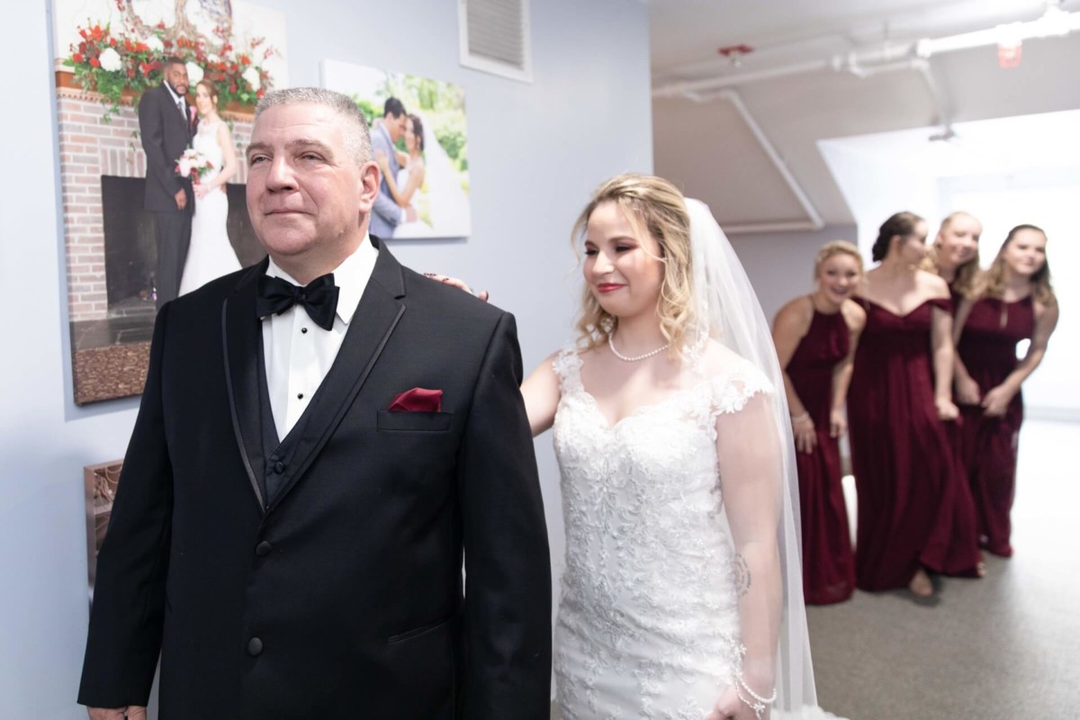 Shannon-James-Wedding-Low-Res-46 - TOMMY GATZ ENTERTAINMENT A bride in a white dress touches a man's shoulder in a tuxedo. In the background, four bridesmaids in burgundy dresses are smiling. Wedding photos hang on the wall.