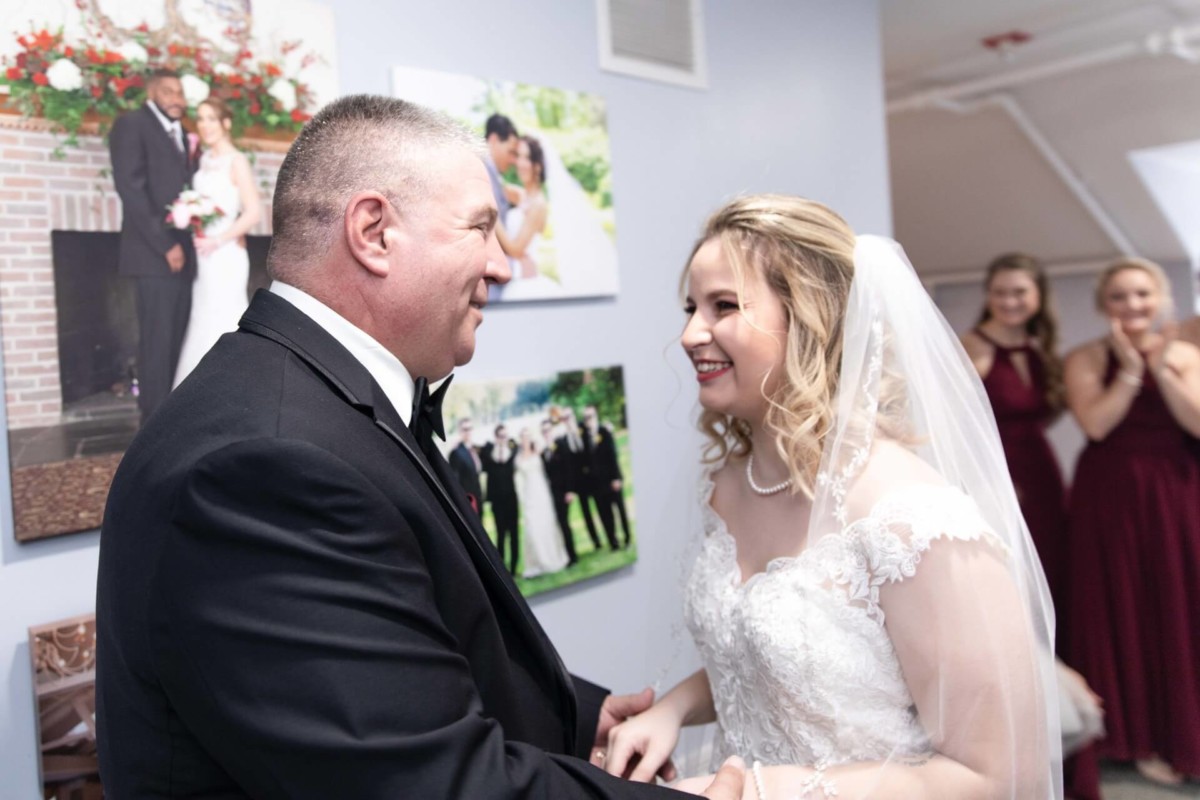 Shannon-James-Wedding-Low-Res-47 - TOMMY GATZ ENTERTAINMENT A bride in a gown and veil smiles at a man in a tuxedo. They are indoors with framed wedding photos on the wall. Bridesmaids are visible in the background.