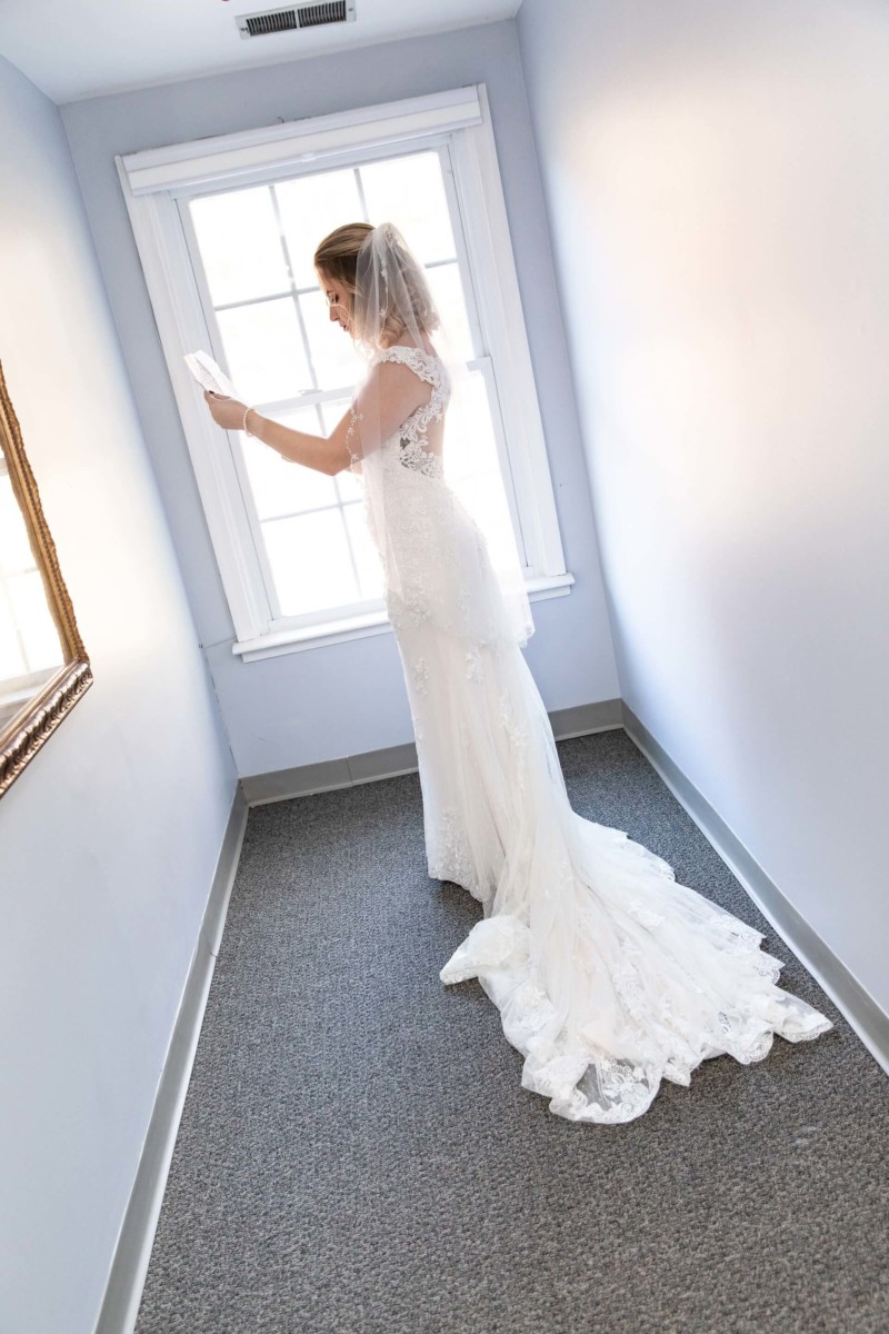 Shannon-James-Wedding-Low-Res-49 - TOMMY GATZ ENTERTAINMENT Bride in a lace wedding dress stands by a window, reading a note, in a narrow, carpeted room.