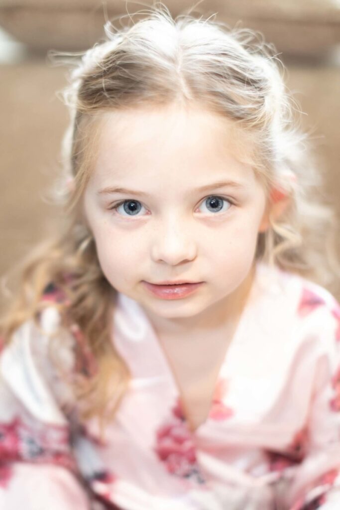 Shannon-James-Wedding-Low-Res-5 - TOMMY GATZ ENTERTAINMENT A young girl with long hair looks directly at the camera, wearing a light pink floral garment.