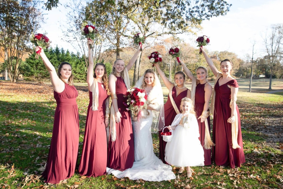 Shannon-James-Wedding-Low-Res-50 - TOMMY GATZ ENTERTAINMENT Bride and seven bridesmaids in matching burgundy dresses pose outdoors with bouquets on an autumn day. Young flower girl in a white dress holds a basket.