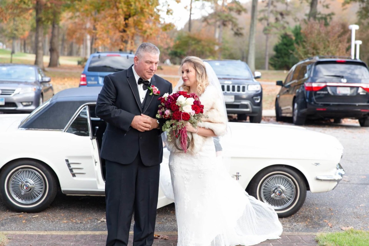 Shannon-James-Wedding-Low-Res-51 - TOMMY GATZ ENTERTAINMENT A bride and a man in formal attire stand by a white vintage car. The bride holds red and white flowers. Trees and parked cars are in the background.