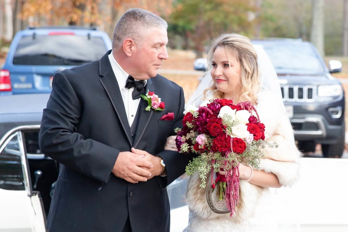 Shannon-James-Wedding-Low-Res-52 - TOMMY GATZ ENTERTAINMENT A bride holding a bouquet stands beside a man in a black suit. They appear to be outdoors, with cars and autumn foliage in the background.