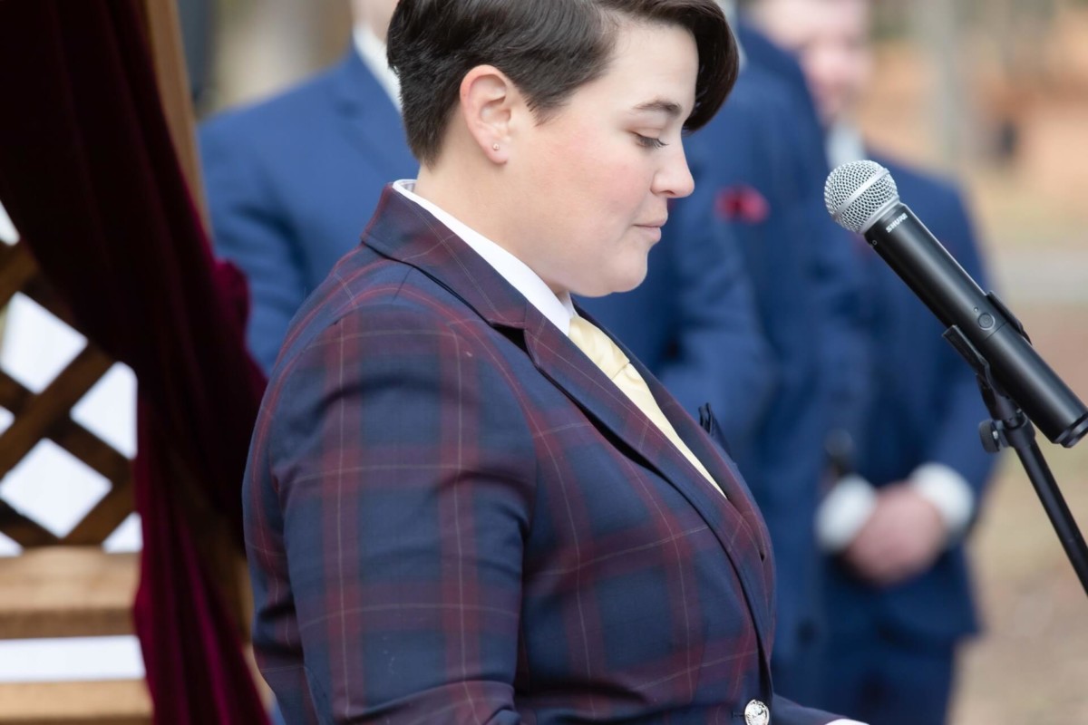 Shannon-James-Wedding-Low-Res-53 - TOMMY GATZ ENTERTAINMENT Person in a plaid suit speaks at a microphone outdoors.