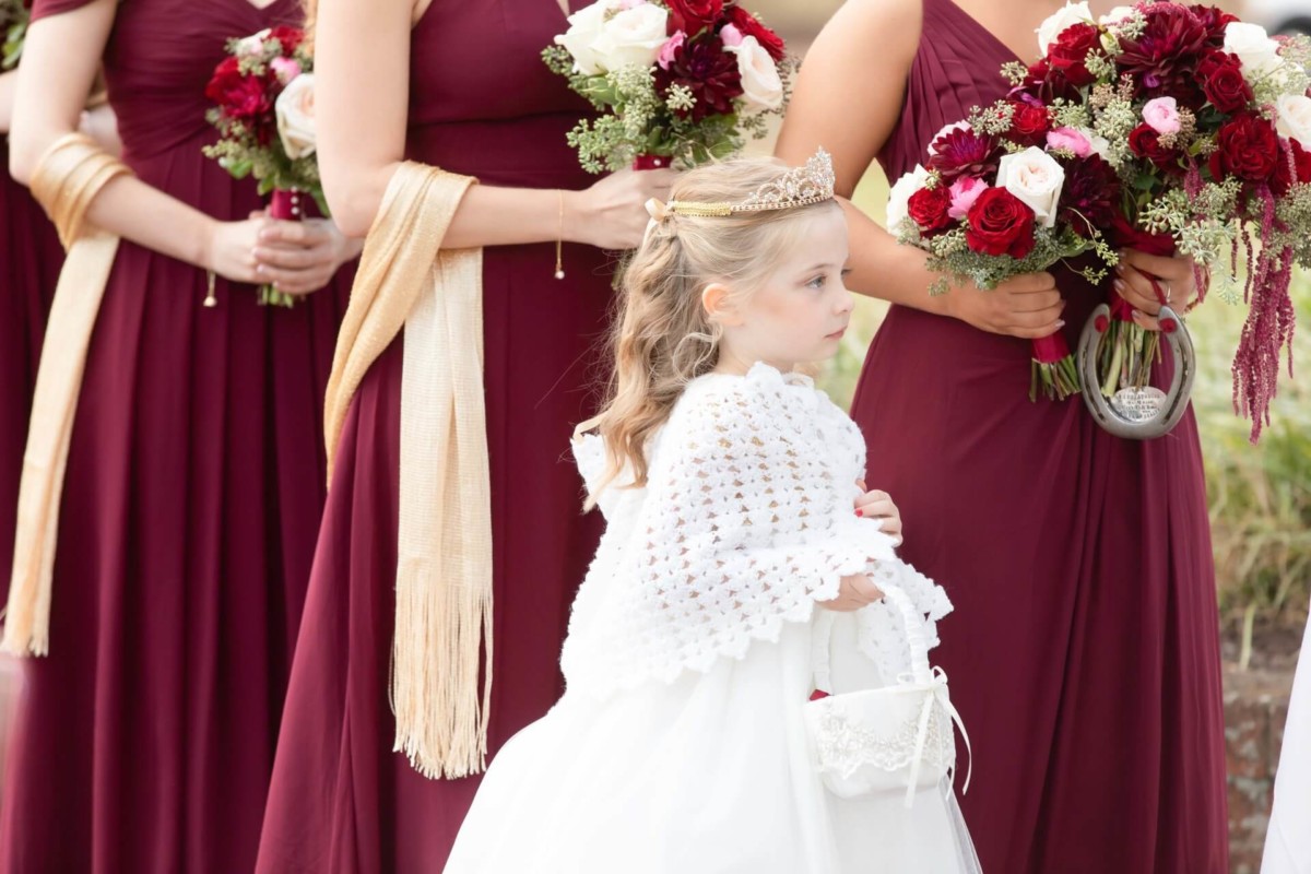 Shannon-James-Wedding-Low-Res-55 - TOMMY GATZ ENTERTAINMENT A young girl in a white dress stands in front of women wearing burgundy gowns and holding bouquets of red and white flowers.