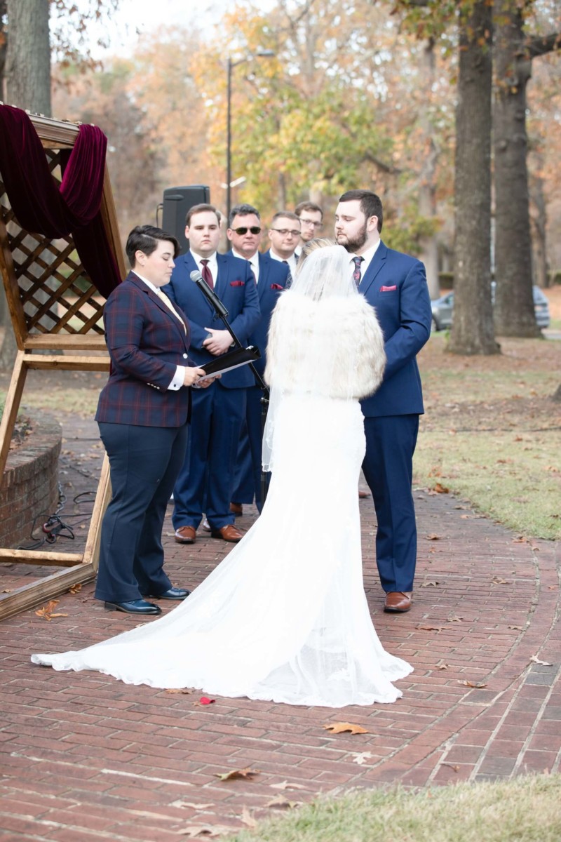 Shannon-James-Wedding-Low-Res-57 - TOMMY GATZ ENTERTAINMENT A bride and groom face each other during an outdoor wedding ceremony. The officiant and groomsmen stand nearby. The scene is surrounded by trees with autumn foliage.