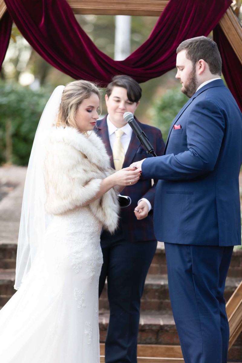 Shannon-James-Wedding-Low-Res-58 - TOMMY GATZ ENTERTAINMENT A bride and groom exchange rings during an outdoor wedding ceremony, with an officiant standing nearby holding a microphone.