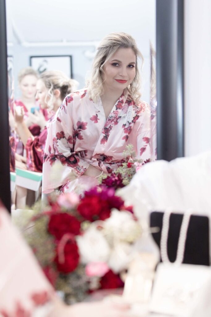 Shannon-James-Wedding-Low-Res-6 - TOMMY GATZ ENTERTAINMENT A woman wearing a floral robe looks at herself in a mirror. A bouquet of red and white flowers is visible in the foreground.