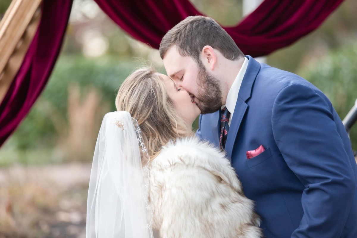 Shannon-James-Wedding-Low-Res-61 - TOMMY GATZ ENTERTAINMENT A couple kissing at an outdoor wedding ceremony. The bride is wearing a fur shawl and veil, and the groom is in a blue suit. The setting includes a burgundy draped arch.