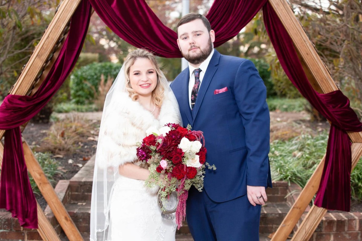 Shannon-James-Wedding-Low-Res-62 - TOMMY GATZ ENTERTAINMENT A bride and groom stand in front of a wooden arch adorned with burgundy drapes. The bride holds a bouquet of red and white flowers, while the groom wears a blue suit.