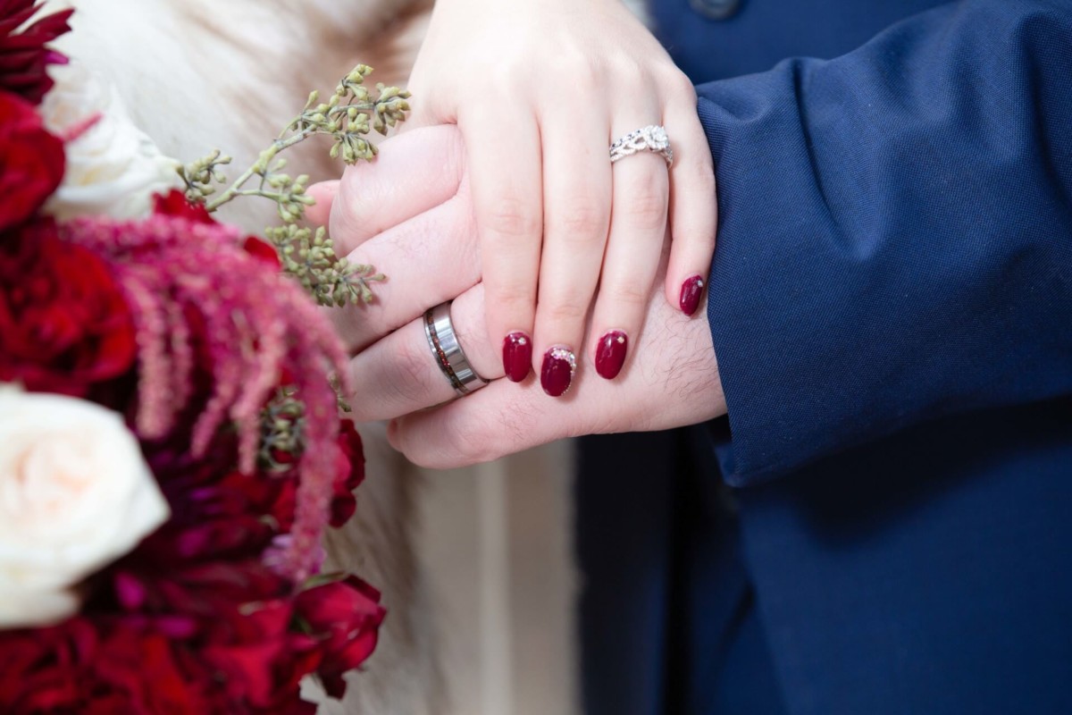 Shannon-James-Wedding-Low-Res-63 - TOMMY GATZ ENTERTAINMENT Two hands with wedding rings are clasped together. One has dark red nail polish and diamond ring. A bouquet of red and white flowers is nearby.