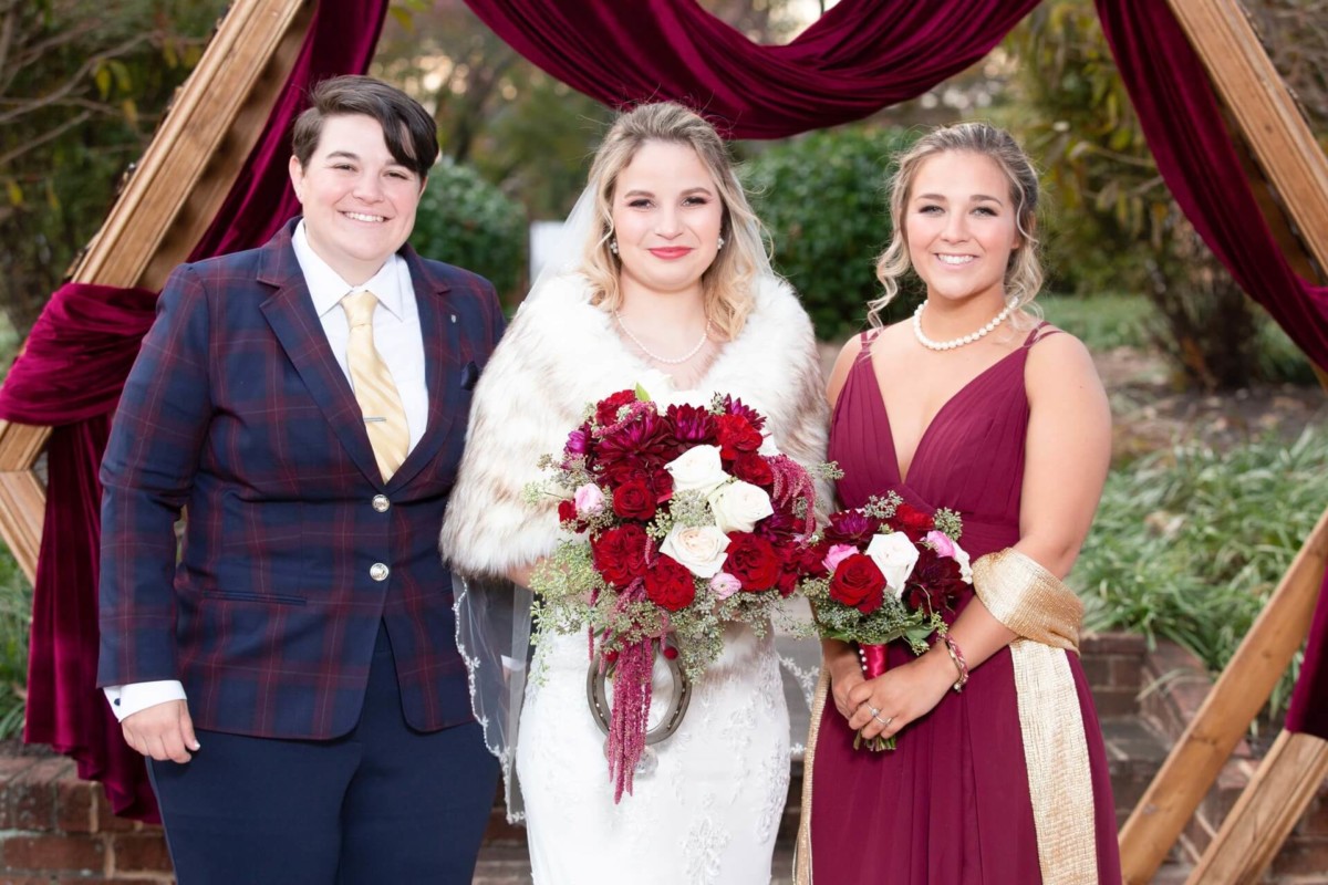 Shannon-James-Wedding-Low-Res-64 - TOMMY GATZ ENTERTAINMENT Three people dressed in formal attire stand outdoors. The center person holds a bouquet of red and white flowers. They are framed by a wooden arch draped with burgundy fabric.