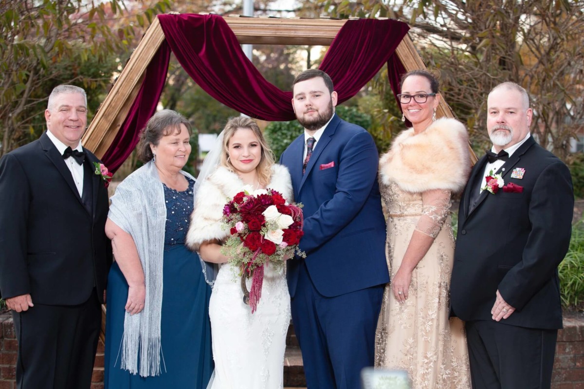 Shannon-James-Wedding-Low-Res-65 - TOMMY GATZ ENTERTAINMENT A wedding group photo with six adults. The bride holds a bouquet, and the groom stands beside her under a wooden arch adorned with red fabric. The others are dressed formally.