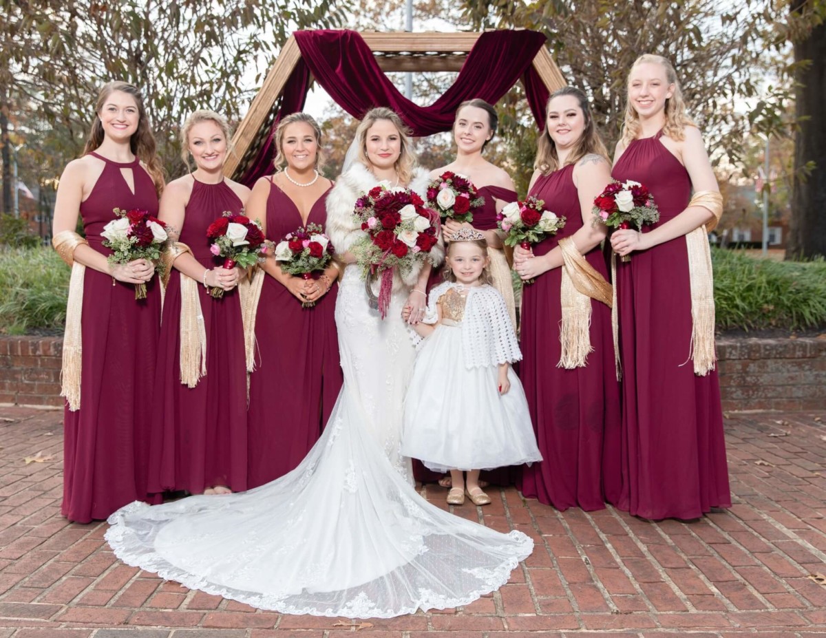 Shannon-James-Wedding-Low-Res-67 - TOMMY GATZ ENTERTAINMENT Bride in a white dress stands with six bridesmaids in burgundy dresses and a flower girl holding bouquets, in front of a wooden arch with drapery outdoors.