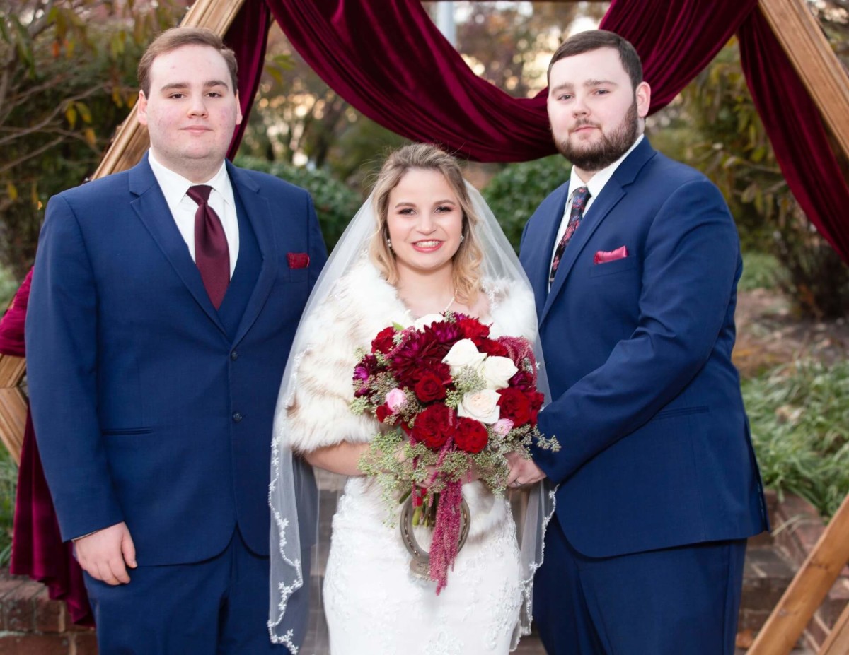 Shannon-James-Wedding-Low-Res-68 - TOMMY GATZ ENTERTAINMENT A bride in a white dress and fur stole stands between two groomsmen in blue suits, holding a red and white bouquet, in front of a burgundy draped arch.
