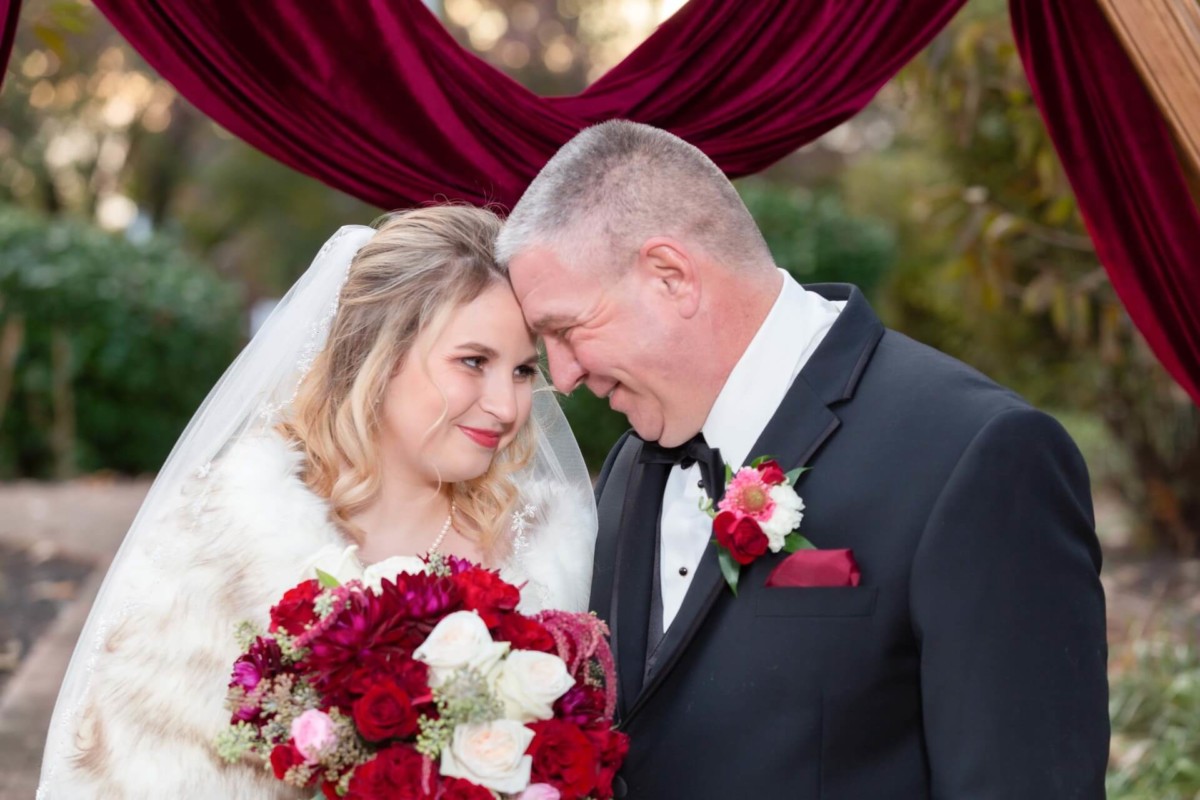 Shannon-James-Wedding-Low-Res-69 - TOMMY GATZ ENTERTAINMENT A couple in formal attire smiles while touching foreheads under a burgundy arch. The woman holds a bouquet of red and white flowers.