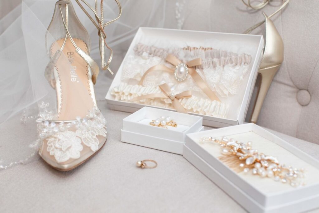 Shannon-James-Wedding-Low-Res-7 - TOMMY GATZ ENTERTAINMENT Bridal accessories including lace shoes, gold heels, a pearl hairpiece, a garter set, earrings, and a ring displayed on a cushioned surface.