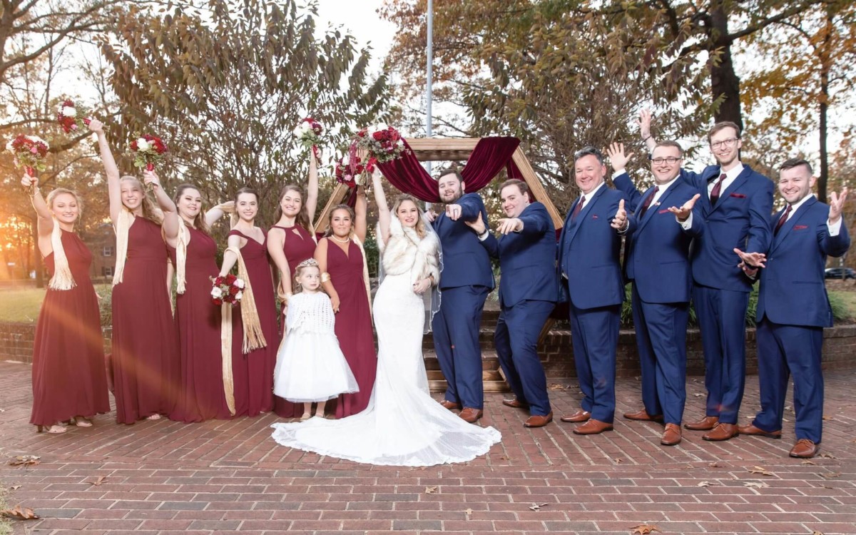 Shannon-James-Wedding-Low-Res-70 - TOMMY GATZ ENTERTAINMENT Wedding party posing outdoors, with bridesmaids in burgundy dresses and groomsmen in blue suits, around a bride in a white gown and a young flower girl in white.
