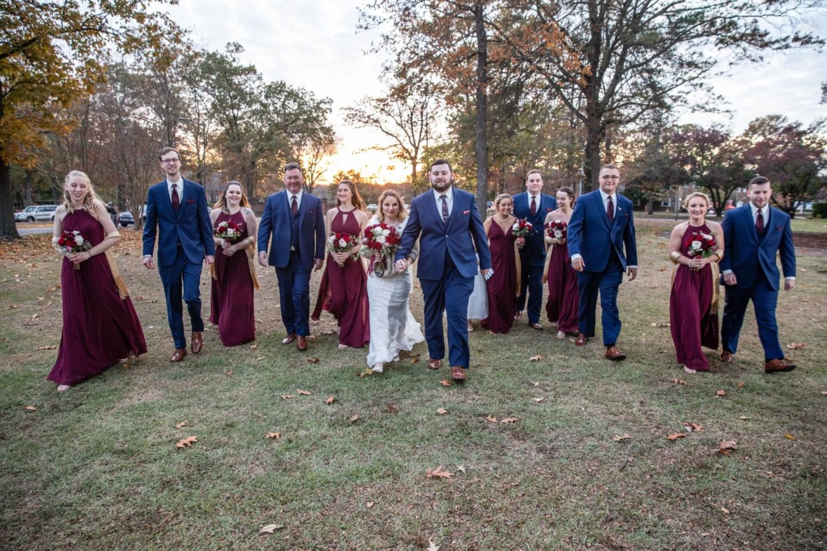 Shannon-James-Wedding-Low-Res-71 - TOMMY GATZ ENTERTAINMENT Wedding party walking across a grassy field, featuring bridesmaids in burgundy dresses and groomsmen in navy suits.