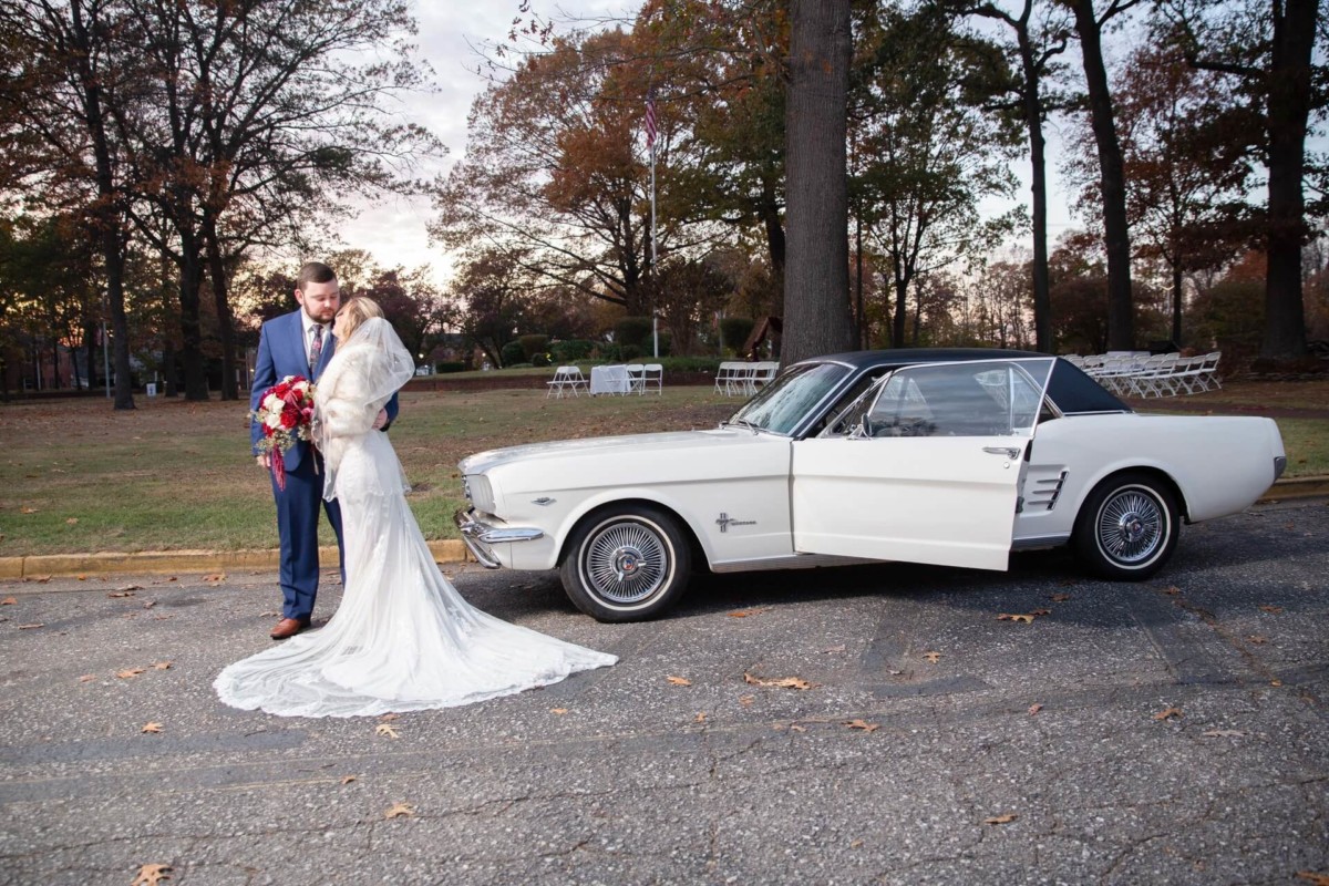 Shannon-James-Wedding-Low-Res-72 - TOMMY GATZ ENTERTAINMENT Bride and groom stand beside a vintage white car in a park, with autumn trees in the background.