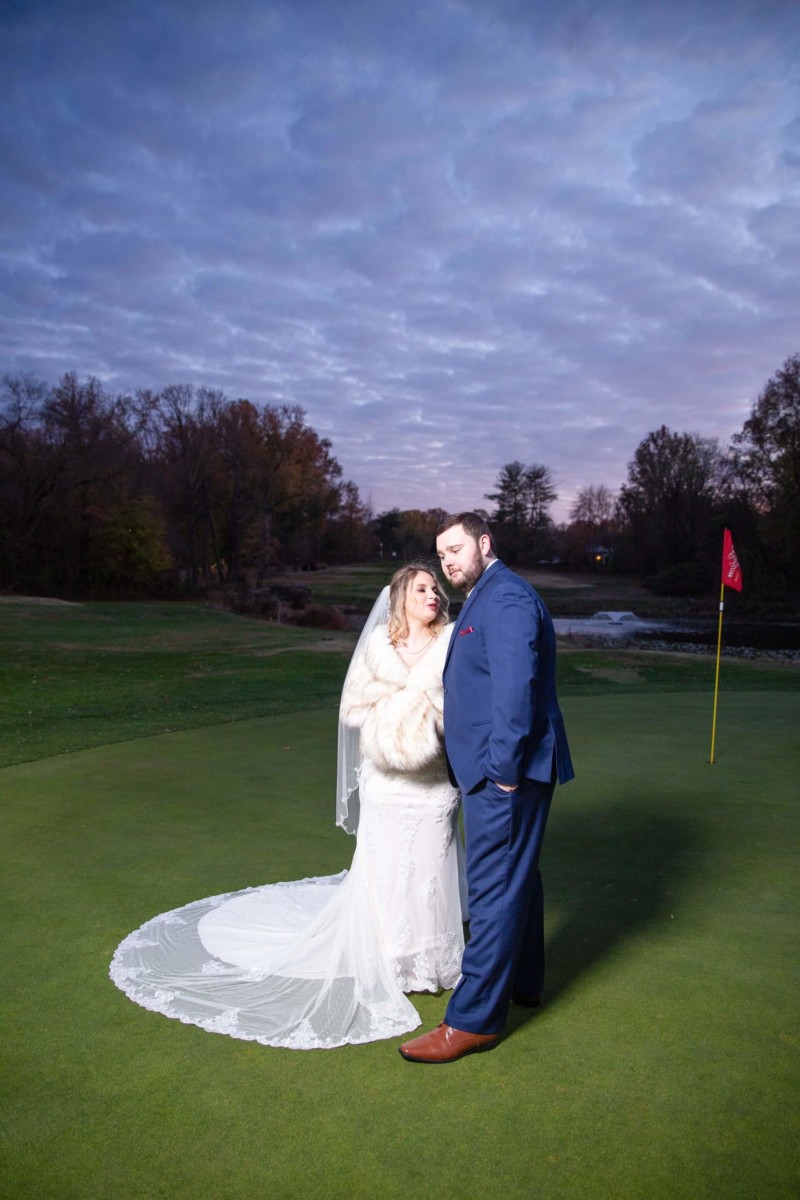 Shannon-James-Wedding-Low-Res-73 - TOMMY GATZ ENTERTAINMENT A bride and groom stand together on a golf course green at dusk, with a red flag in the background. The bride wears a white dress and veil, and the groom is in a blue suit.
