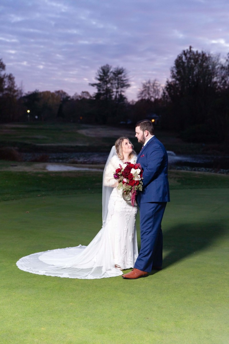 Shannon-James-Wedding-Low-Res-74 - TOMMY GATZ ENTERTAINMENT A bride and groom stand together on a grassy field at dusk, with the bride holding a bouquet of red and white flowers.