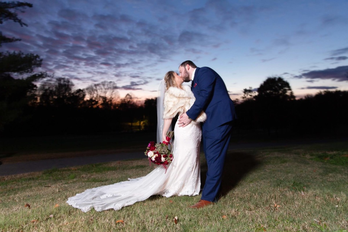 Shannon-James-Wedding-Low-Res-75 - TOMMY GATZ ENTERTAINMENT A couple in wedding attire shares a kiss outdoors at dusk, with a dramatic sky and silhouetted trees in the background.