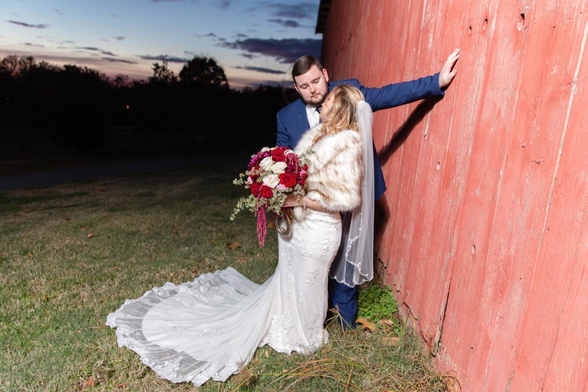 Shannon-James-Wedding-Low-Res-76 - TOMMY GATZ ENTERTAINMENT A bride and groom pose together against a red barn at sunset. The bride holds a bouquet of red and white flowers and wears a lace gown with a fur shawl; the groom is in a blue suit.