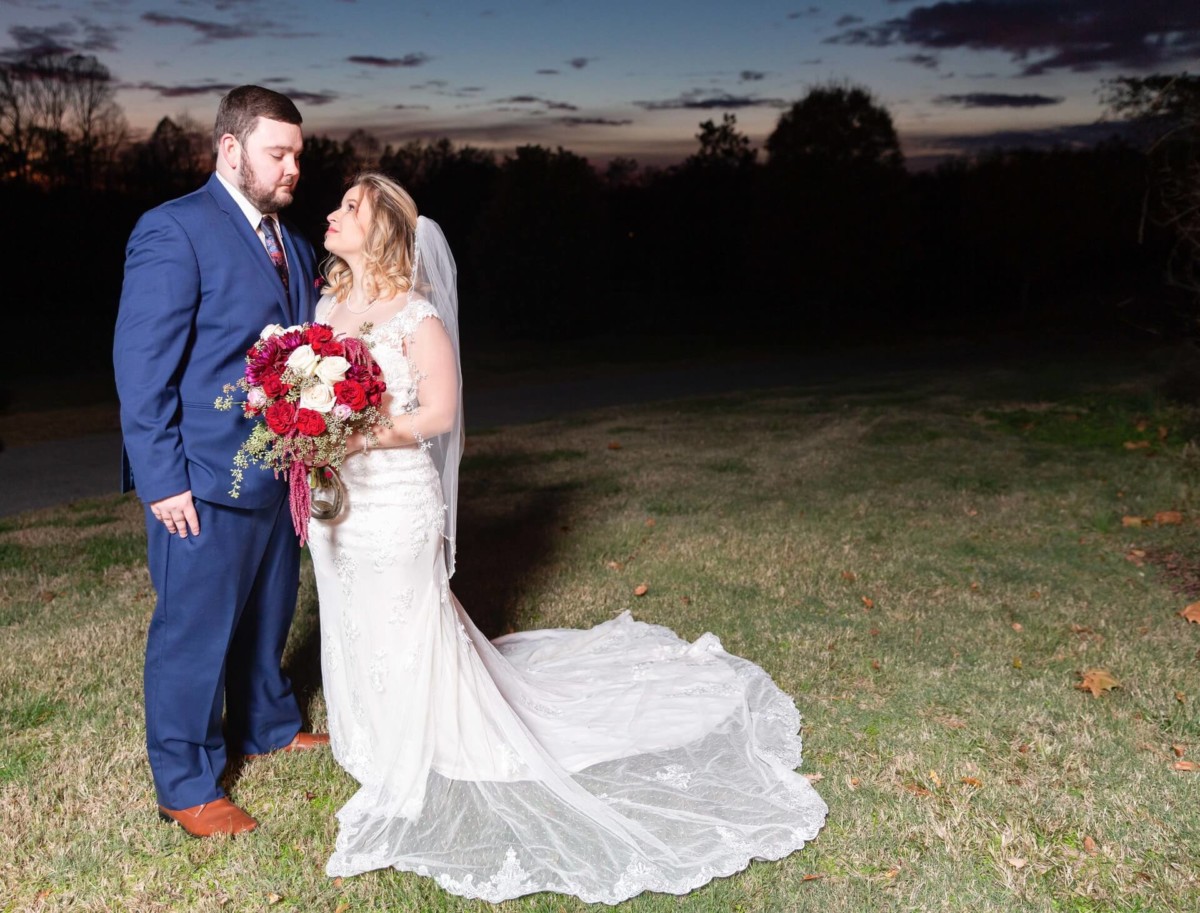 Shannon-James-Wedding-Low-Res-77 - TOMMY GATZ ENTERTAINMENT Bride and groom standing outdoors at dusk, with the bride holding a bouquet of red and white flowers.