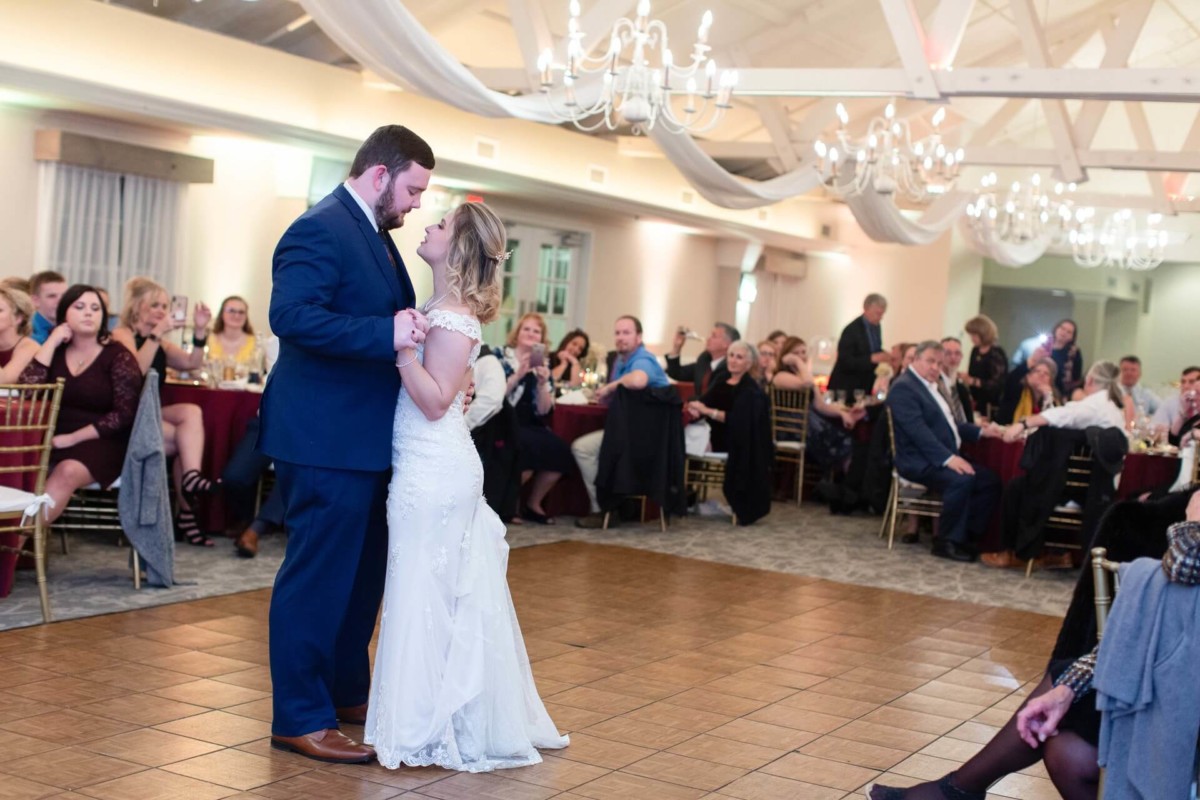 Shannon-James-Wedding-Low-Res-78 - TOMMY GATZ ENTERTAINMENT A bride and groom dance together in a ballroom with guests seated at tables in the background. Chandeliers and ceiling drapery are visible.
