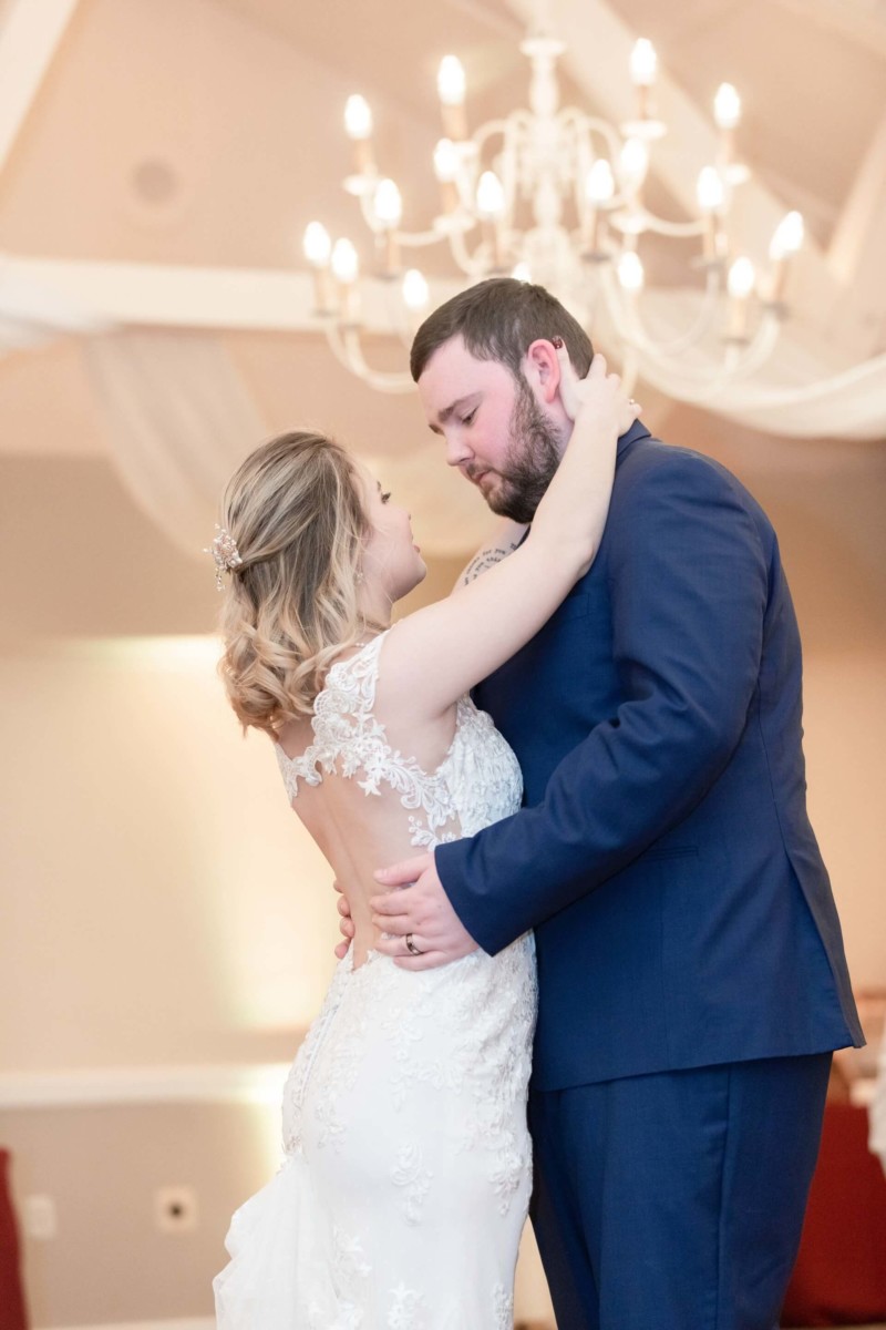 Shannon-James-Wedding-Low-Res-80 - TOMMY GATZ ENTERTAINMENT A bride and groom share a dance in a warmly lit room, with a chandelier visible in the background. The bride wears a white lace gown, and the groom is in a blue suit.