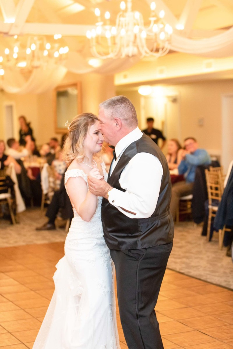 Shannon-James-Wedding-Low-Res-83 - TOMMY GATZ ENTERTAINMENT A bride and an older man, both in formal attire, share a dance in a warmly lit reception hall with guests seated in the background.