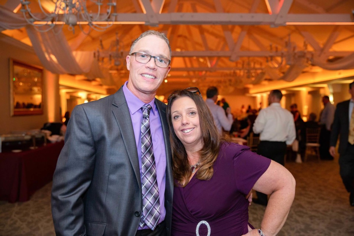 Shannon-James-Wedding-Low-Res-84 - TOMMY GATZ ENTERTAINMENT A man and a woman dressed in formal attire pose together, smiling in a warmly lit event venue with a visible ceiling and partygoers in the background.