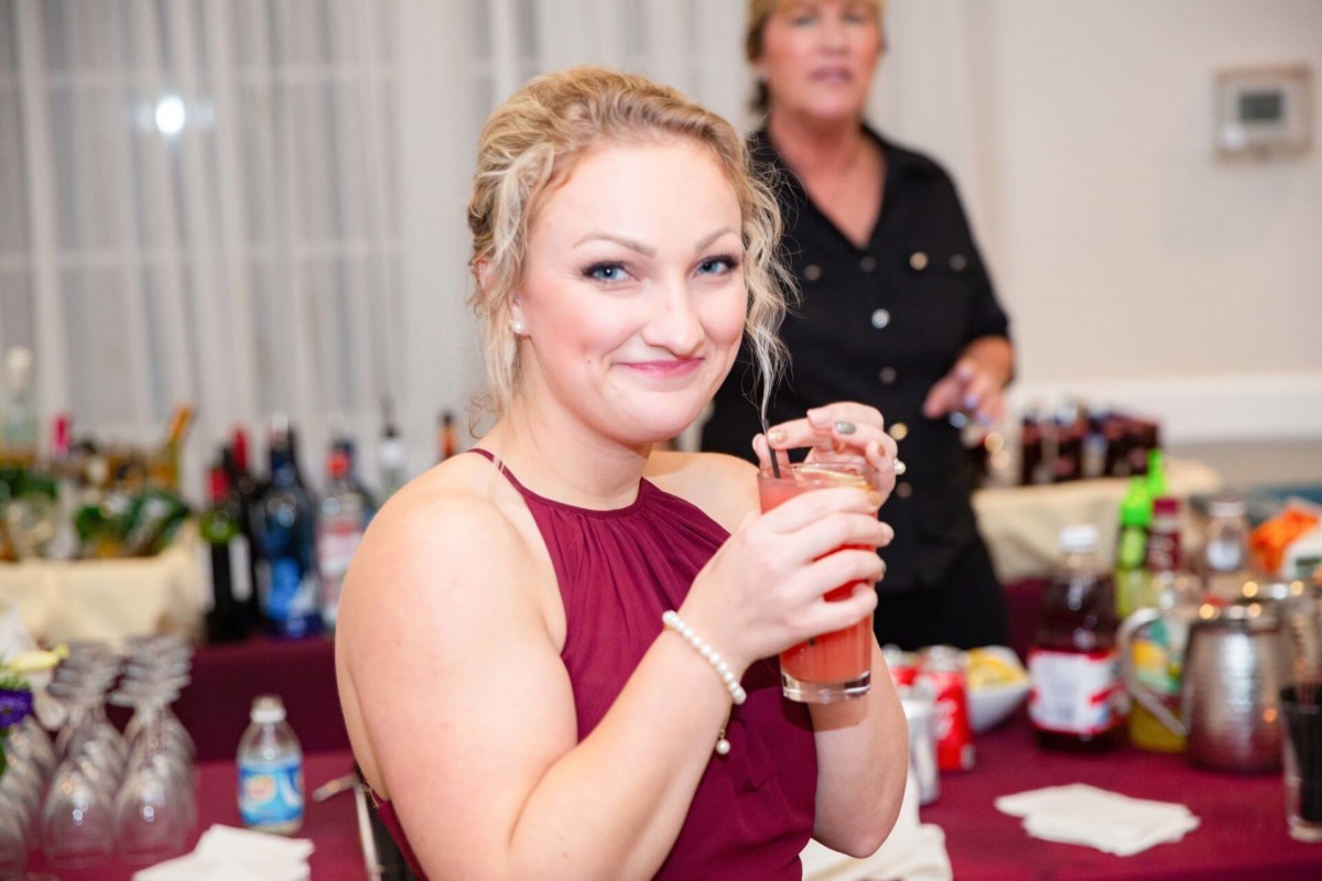 Shannon-James-Wedding-Low-Res-85 - TOMMY GATZ ENTERTAINMENT A person in a sleeveless red dress holds a drink and smiles at the camera, with a table of beverages and a second person in the background.