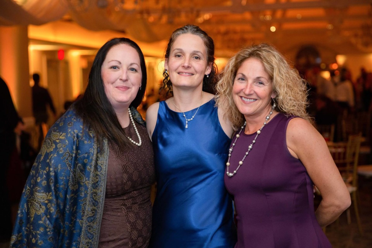 Shannon-James-Wedding-Low-Res-86 - TOMMY GATZ ENTERTAINMENT Three women standing together indoors, smiling at the camera, dressed in formal attire with necklaces.