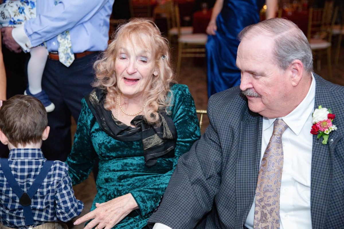 Shannon-James-Wedding-Low-Res-87 - TOMMY GATZ ENTERTAINMENT An elderly couple sits together at an event. The woman wears a green dress, and the man is in a suit with a floral boutonniere. A child in a plaid shirt is in the foreground.