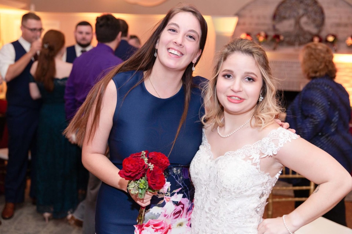 Shannon-James-Wedding-Low-Res-88 - TOMMY GATZ ENTERTAINMENT Two women smile at a wedding reception; one holds a bouquet. Guests are seen in the background.