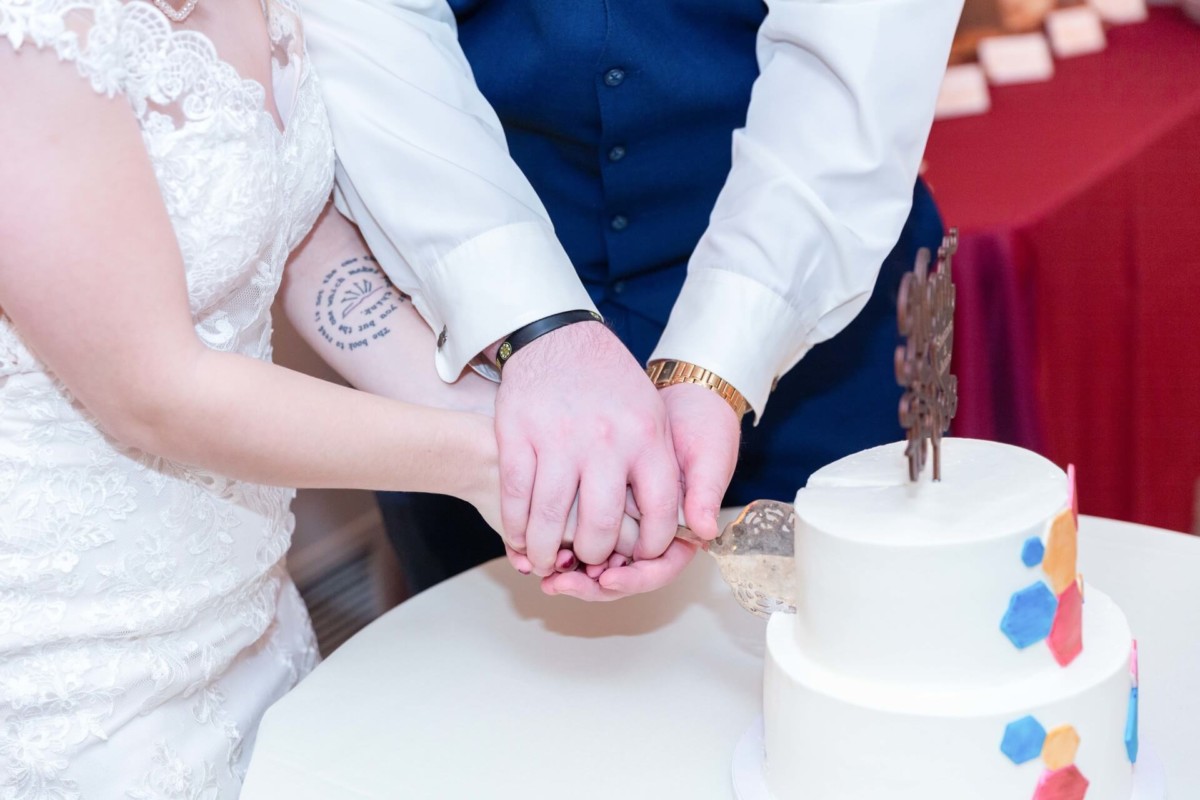 Shannon-James-Wedding-Low-Res-89 - TOMMY GATZ ENTERTAINMENT A couple cuts a white two-tier cake together. One person wears a lace dress, the other a navy vest. The cake features colorful hexagonal decorations and a chocolate topper.