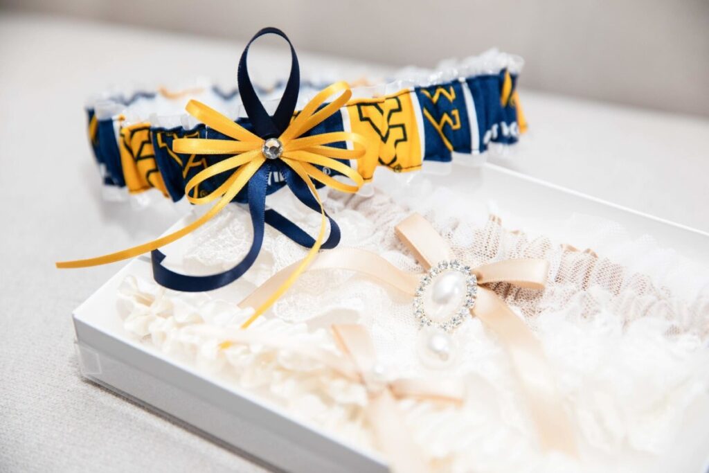 Shannon-James-Wedding-Low-Res-9 - TOMMY GATZ ENTERTAINMENT A close-up of two wedding garters: one with blue and yellow ribbons, the other with lace and a pearl embellishment, displayed in a white box.