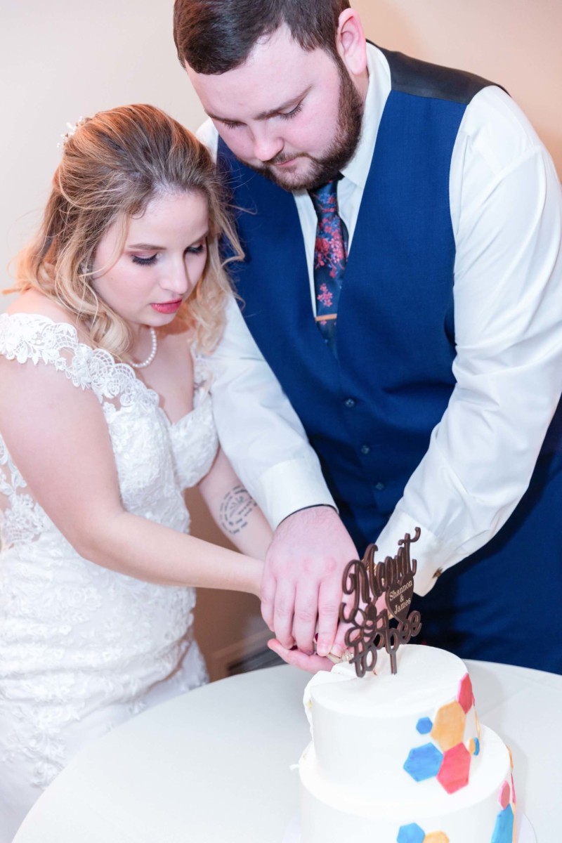 Shannon-James-Wedding-Low-Res-90 - TOMMY GATZ ENTERTAINMENT A bride and groom cut a white wedding cake decorated with colorful hexagons and a "Mr. & Mrs." topper.