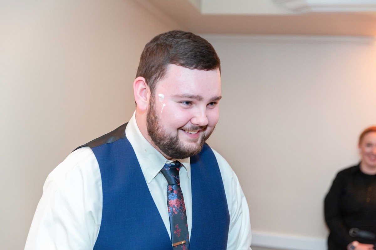 Shannon-James-Wedding-Low-Res-92 - TOMMY GATZ ENTERTAINMENT A man with a beard smiling, wearing a blue vest and tie, stands in a room with white walls. A woman in black is visible in the background.