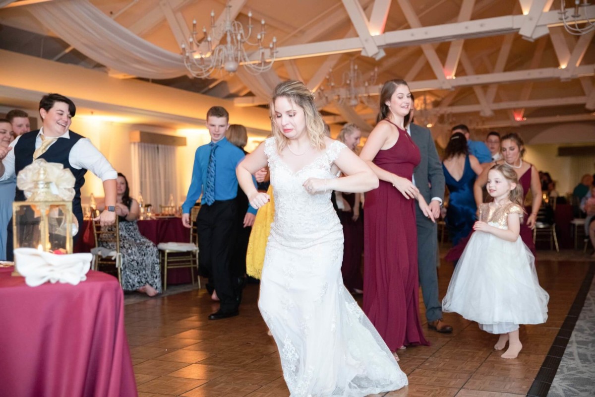 Shannon-James-Wedding-Low-Res-93 - TOMMY GATZ ENTERTAINMENT A bride in a white dress leads a group of people, including a girl in a white dress, dancing in a well-lit room with chandeliers and wood beams.