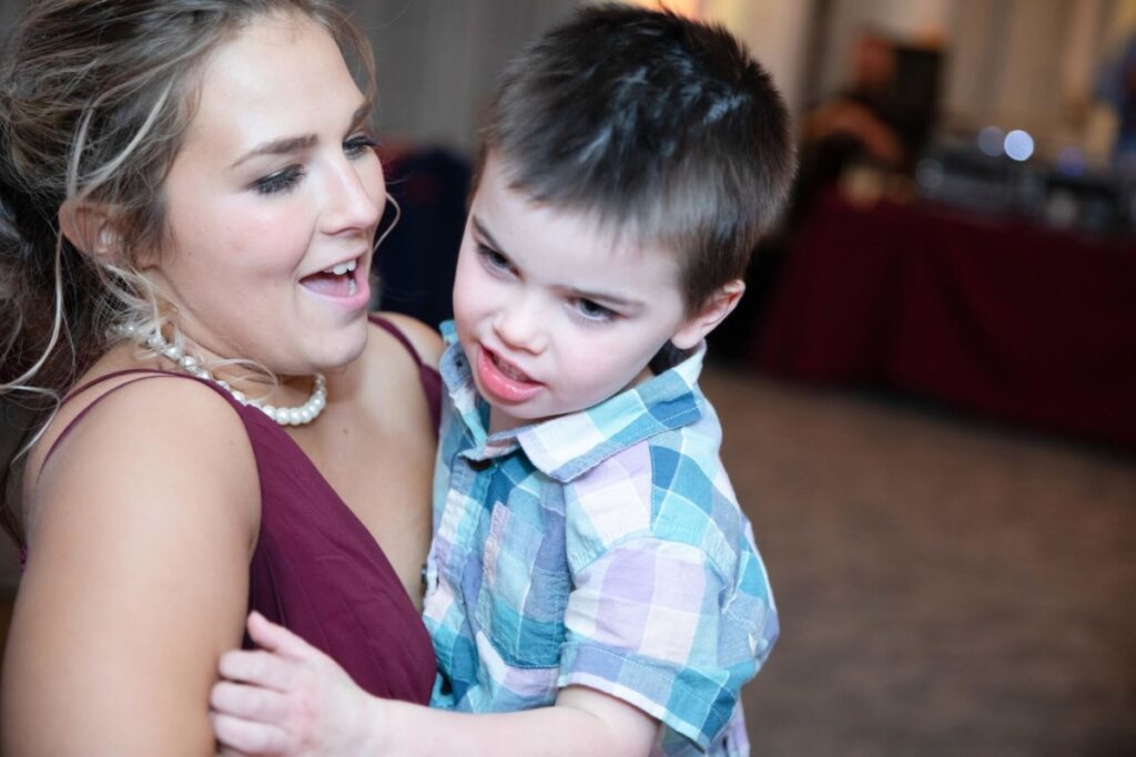 Shannon-James-Wedding-Low-Res-94 - TOMMY GATZ ENTERTAINMENT A woman holding a young boy. Both appear to be engaged in a lively interaction. The woman is wearing a burgundy top and a pearl necklace, while the boy is in a plaid shirt.