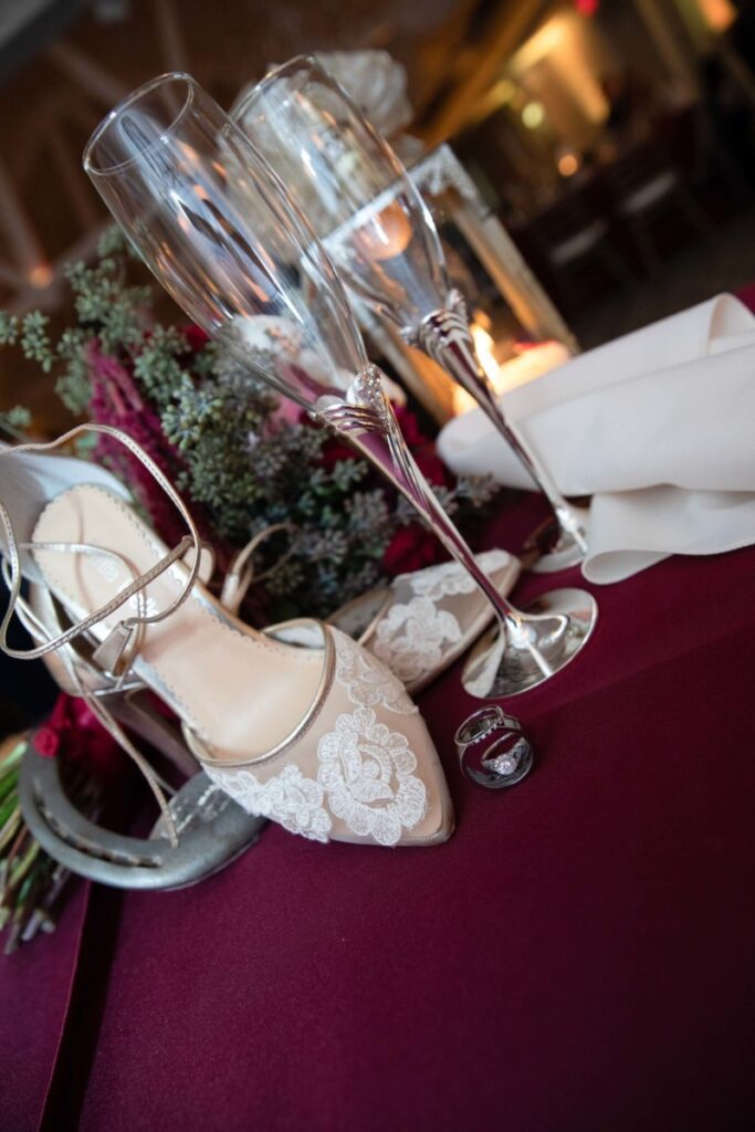 Shannon-James-Wedding-Low-Res-95 - TOMMY GATZ ENTERTAINMENT Wedding still life with lace high-heeled shoes, two champagne flutes, and rings on a burgundy tablecloth.